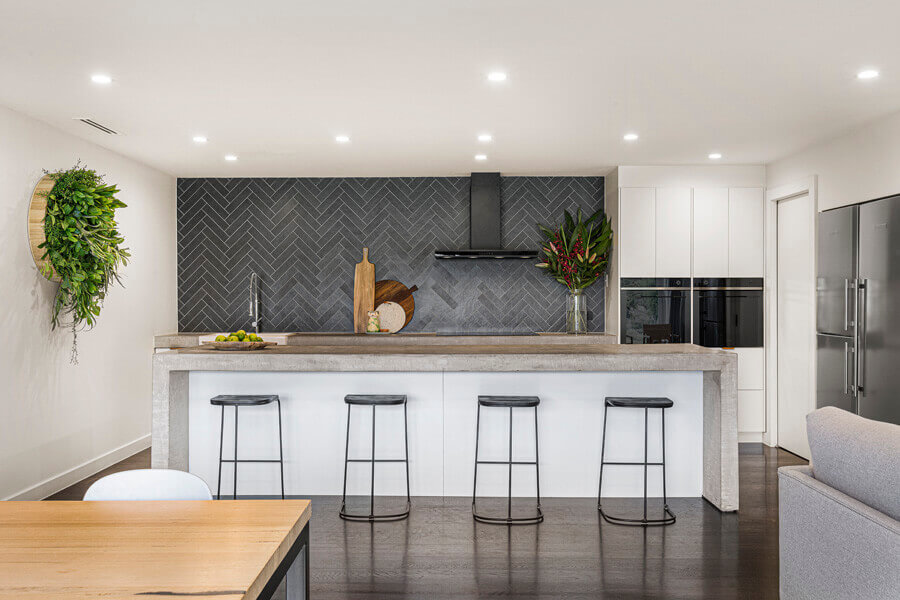 Handleless white kitchen with a charcoal herringbone tiled wall, concrete-topped breakfast bar and black stools